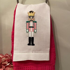 May include: White hand towel with an embroidered pink and green nutcracker design. The towel is hanging on a red towel rack.