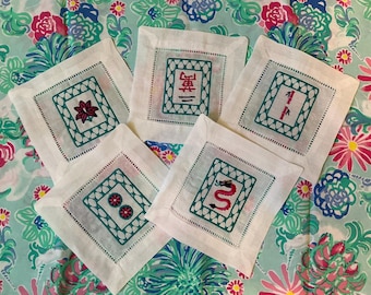May include: A collection of five white linen coasters with embroidered designs. Each coaster features a square design with a teal border and a central image in red and teal. The images include a flower, the number one, two circles, a dragon, and a Chinese character.
