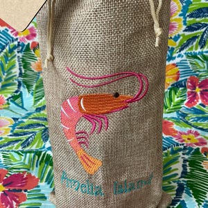 May include: A beige burlap drawstring bag with an embroidered shrimp design in shades of pink and orange. The text "Amelia Island" is embroidered in teal. The bag is on a colorful floral patterned surface.