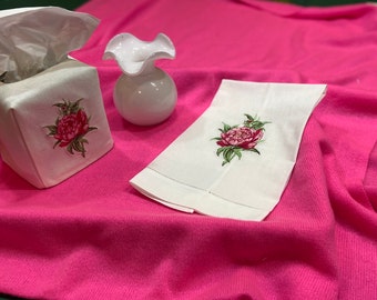 May include: A white tissue box cover, a small white vase, and a white hand towel are displayed on a pink surface. Each item features a detailed embroidered pink peony with green leaves. The tissue box is square, and the vase has a ruffled top.
