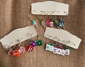 May include: A collection of wine glass charms featuring various designs including miniature pickleball paddles, colorful translucent dice, and white mahjong tiles. Each charm is attached to a metal hoop accented with small, multi-colored beads.