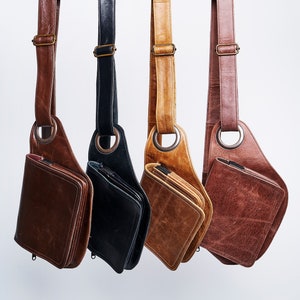 Könnte beinhalten: Vier Leder-Umhängetaschen hängen an einem schwarzen Metallgestell. Die Taschen sind braun, beige, schwarz und dunkelbraun. Jede Tasche hat einen einzigen Riemen und einen Reißverschluss.