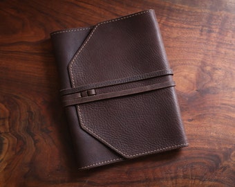 May include: A dark brown leather journal with a textured surface and a strap closure. The journal has a geometric design on the cover and is stitched with white thread. The background is a wooden surface.