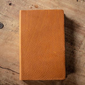 May include: A rectangular notebook with a textured, tan-colored cover. The surface of the cover displays a distinct, pebbled grain pattern across the entire front face, showing a consistent warm brown tone throughout the material.