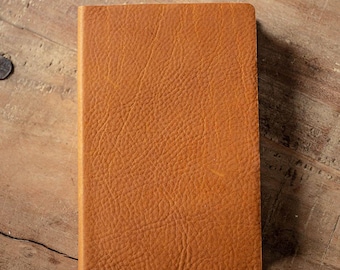 May include: A rectangular notebook with a textured, tan-colored cover. The surface of the cover displays a distinct, pebbled grain pattern across the entire front face, showing a consistent warm brown tone throughout the material.