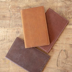 May include: Four leather journals in shades of brown, tan, and dark brown are arranged on a wooden surface. The journals are rectangular with stitched edges, suggesting a handcrafted design. The varying colors and textures create a rustic aesthetic.