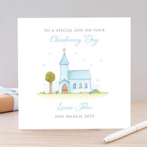 Op de afbeelding: Een vierkante wenskaart met een aquarelillustratie van een lichtblauwe kerk met een bruine deur, een groene boom en gras. De tekst luidt: TO A SPECIAL SON ON YOUR Christening Day, Lucas John, en 2ND MARCH 2025 in grijs en blauw.