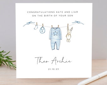 Puede incluir: Tarjeta de felicitación blanca con una ilustración en acuarela de ropa de bebé colgada en un tendedero. El texto dice "CONGRATULATIONS KATE AND LIAM ON THE BIRTH OF YOUR SON" y "Theo Archie 21.10.23". Un bolígrafo descansa sobre la superficie de madera.
