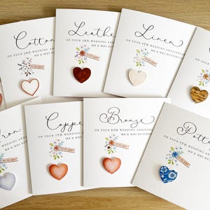 May include: A selection of white anniversary cards, each with a different theme. The cards are adorned with a heart and floral design. The cards are labelled with anniversary milestones such as "Paper", "Cotton", "Leather", and "Linen".