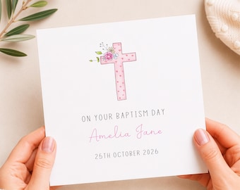 May include: A square white greeting card featuring a pink cross decorated with small hearts and a floral cluster. The text reads ON YOUR BAPTISM DAY, Amelia Jane, and 25TH OCTOBER 2026 in black and pink lettering centred on the card.