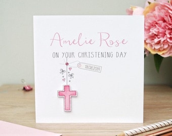 May include: A square white greeting card for a christening featuring the name Amelie Rose in pink script and the text ON YOUR CHRISTENING DAY in black. A pink cross illustration hangs from a string with small bows, hearts, and a date tag 18.08.2019.