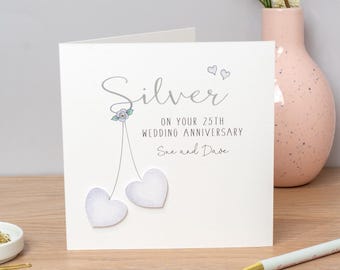 May include: A white square greeting card featuring the text Silver ON YOUR 25TH WEDDING ANNIVERSARY Sue and Dave. The design includes two light gray heart-shaped embellishments hanging from thin lines, a small floral detail, and two tiny hearts.