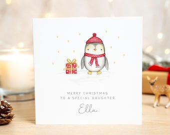 Könnte beinhalten: Eine quadratische weiße Grußkarte mit einem gezeichneten Pinguin in roter Mütze und Schal, der neben einem roten Geschenk mit goldener Schleife steht. Der Text lautet MERRY CHRISTMAS TO A SPECIAL DAUGHTER und der Name Ella in Schreibschrift.