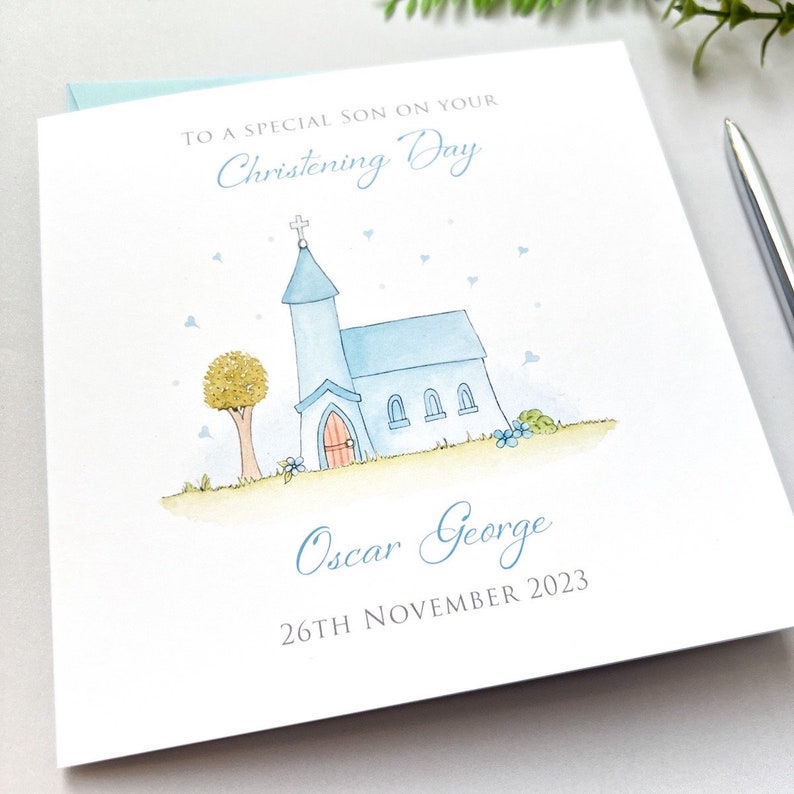 Op de afbeelding: Een vierkante witte wenskaart met een aquarelillustratie van een lichtblauwe kerk met een kruis, een boom en kleine blauwe hartjes. De tekst luidt: TO A SPECIAL SON ON YOUR Christening Day, Oscar George, 26TH NOVEMBER 2023.