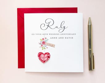 Peut inclure: Une carte de vœux carrée blanche avec une enveloppe rouge et un stylo doré. La carte présente le texte Ruby, ON YOUR 40TH WEDDING ANNIVERSARY, ANNE AND DAVID, une illustration aquarelle d'une bannière florale avec 14.04.24 et un cœur rouge facetté.