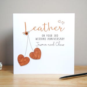 May include: A white square greeting card with the text Leather ON YOUR 3RD WEDDING ANNIVERSARY Jasmin and Chris. The design features a copper-coloured script, a small orange flower, and two dangling brown heart shapes resembling cherries.