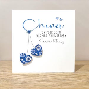 May include: A square white greeting card featuring the blue text China ON YOUR 20TH WEDDING ANNIVERSARY and the names Anna and Terry. Two blue and white floral patterned heart-shaped embellishments hang from a blue floral illustration.
