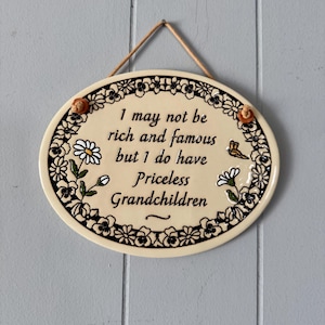 May include: Cream oval ceramic plaque with the text "I may not be rich and famous but I do have Priceless Grandchildren." It features a black floral border and a brown rope for hanging. The plaque is a decorative item.