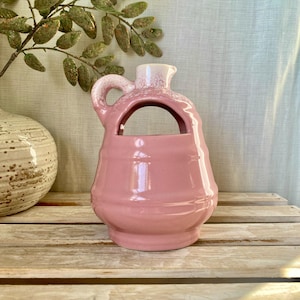 May include: A pink ceramic pitcher with a handle and a unique opening. The pitcher has a light pink body and a white top. The pitcher is sitting on a wooden surface.