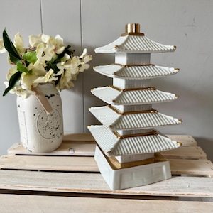 May include: A white ceramic pagoda with gold detailing, featuring multiple tiered roofs. Next to it, a distressed white mason jar vase holds a bouquet of cream-coloured flowers. The items are displayed on a white wooden crate.