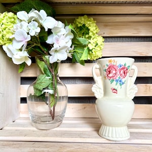 May include: A clear glass vase holds white and light green flowers. A cream-colored vase with two handles and floral designs sits to the right. The vases are displayed on a wooden crate.