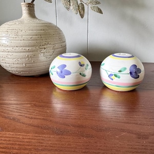 May include: Two spherical ceramic salt and pepper shakers with a floral pattern. Each shaker has a blue flower, green leaves, and horizontal stripes of yellow, pink, and green. A textured, beige vase is in the background.