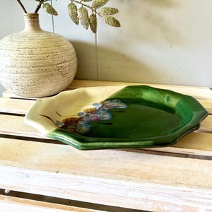 May include: A ceramic plate with a scalloped edge featuring a two-tone glaze. The left side is a creamy off-white, while the right side is a deep, glossy forest green. A small cluster of blue and brown floral-like painted accents sits in the center.