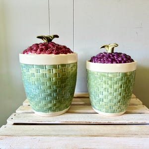 May include: Two ceramic canisters with a light green basket-weave texture on the base and a cream-colored rim. The lids are shaped like clusters of fruit, with one featuring deep red berries and the other featuring dark purple grapes.