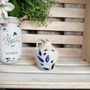 May include: A small, off-white ceramic pitcher with a blue leaf design. The pitcher has a handle and several small holes near the top. It is displayed on a wooden shelf with a mason jar and greenery.