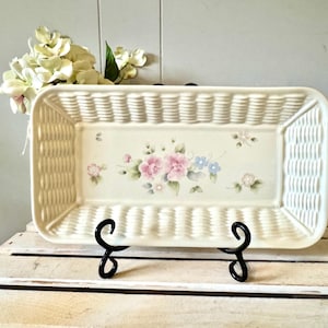 May include: A rectangular off-white ceramic basket-weave tray featuring a central floral motif with pink and blue flowers and green leaves. The tray is displayed on a black metal stand, showcasing a textured exterior and a smooth interior base.