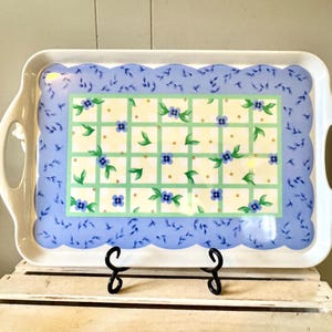 May include: A rectangular white serving tray with integrated handles featuring a central grid pattern of light green lines, small yellow dots, and blue floral accents, surrounded by a scalloped border of light blue with dark blue leaf motifs.