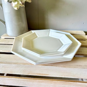 May include: Two stacked, off-white octagonal ceramic plates with a smooth, glossy finish. The plates feature a distinct geometric eight-sided shape with raised edges, creating a clean and uniform appearance for this set of dinnerware.