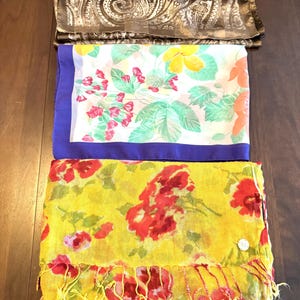 May include: Three folded scarves. The top scarf is brown with a paisley pattern. The middle scarf is white with a floral pattern and a blue border. The bottom scarf is yellow with red flowers and fringe.
