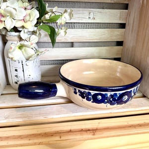 May include: A ceramic serving bowl with a handle, featuring a cream-colored interior and a navy blue rim and handle. The bowl is decorated with blue floral patterns. The bowl is displayed on a wooden crate with a vase of white flowers.