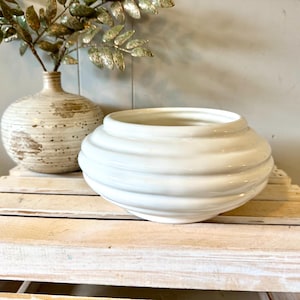 May include: A glossy white ceramic bowl with a rounded, ribbed silhouette and a wide opening. The bowl features a smooth, reflective surface with horizontal ridges circling the exterior, creating a layered, bulbous shape that tapers towards the base.