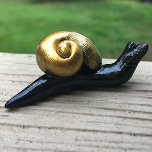 May include: A handcrafted snail figurine with a glossy black body and a golden, spiraled shell. The snail is positioned on a light-colored surface, with a blurred green background. The sculpture is detailed and appears to be made of a durable material.