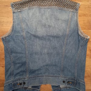 May include: A blue denim vest with silver studs on the back. The vest has a classic denim jacket style with two pockets on the front and a button closure.