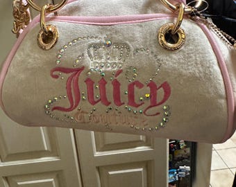 May include: Cream-colored velour handbag with pink trim and handles. The bag features the word "Juicy" in pink script, embellished with rhinestones and a crown design. Gold-tone hardware and a chain accent complete the design.
