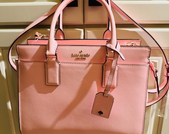 May include: A pink leather satchel handbag with a gold logo that reads "kate spade new york". The bag has a top handle and a long shoulder strap.