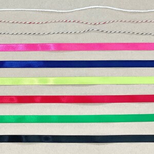 K&ouml;nnte beinhalten: Sortiment aus Band und Schnur in verschiedenen Farben, darunter Wei&szlig;, Rot, Schwarz, Pink, Blau, Gr&uuml;n und Gelb. Das Band und die Schnur sind in Reihen auf einer braunen Oberfl&auml;che angeordnet.