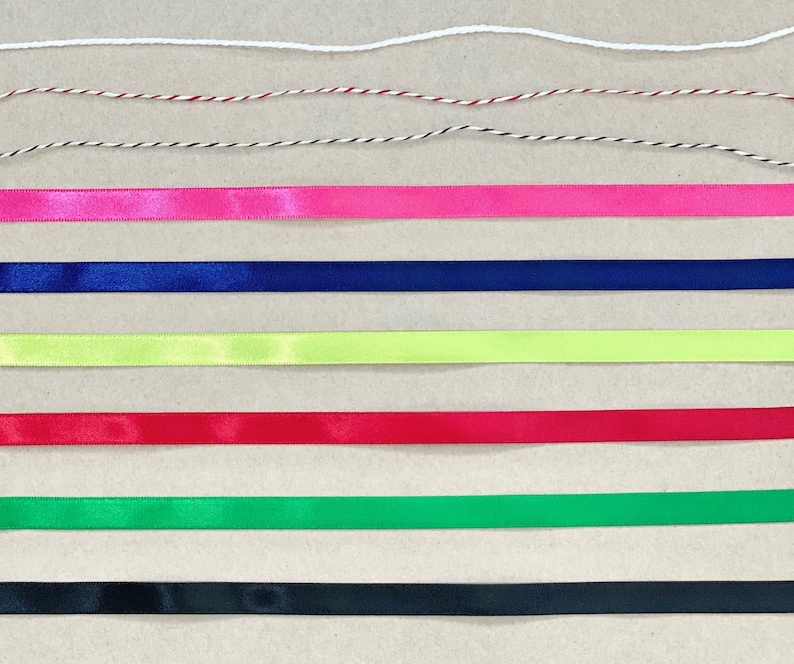 K&ouml;nnte beinhalten: Sortiment aus Band und Schnur in verschiedenen Farben, darunter Wei&szlig;, Rot, Schwarz, Pink, Blau, Gr&uuml;n und Gelb. Das Band und die Schnur sind in Reihen auf einer braunen Oberfl&auml;che angeordnet.