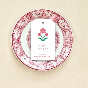 K&ouml;nnte beinhalten: Ein wei&szlig;er Geschenkanh&auml;nger mit einer roten Blumenillustration und dem Text "a gift for you with love, THE SCHAEFERS".