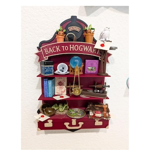 Może przedstawiać: Bordowa półka z wieloma poziomami, ozdobiona miniaturowymi przedmiotami z motywem Harry'ego Pottera. Na górze znajduje się wzór pociągu ze słowami "Express" i "Back to Hogwarts". Przedmioty obejmują książki, zestaw do herbaty i model sowy.