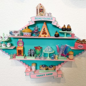 May include: A teal and pink wall-mounted display shelf labeled HONEYDUKES SWEET SHOP, featuring multiple tiers filled with miniature candy-themed items, including small jars, boxes, tiered stands, and various colorful confectionery replicas.
