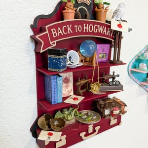 May include: A burgundy-colored, multi-tiered shelf unit with a "Back to Hogwarts" banner. The shelves display miniature items like a tea tin, books, a crystal ball, a necklace, and a model dragon. The top shelf features a train and potted plants.
