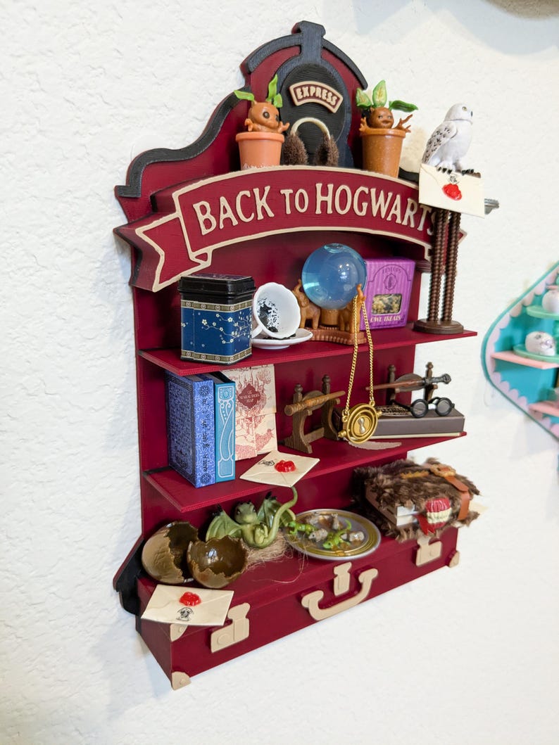 May include: A burgundy-colored, multi-tiered shelf unit with a "Back to Hogwarts" banner. The shelves display miniature items like a tea tin, books, a crystal ball, a necklace, and a model dragon. The top shelf features a train and potted plants.