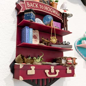 May include: A burgundy-colored shelf unit with three shelves, shaped like a train. The top banner reads "BACK TO HOGWARTS". The shelves display miniature books, a blue globe, a golden necklace, and other themed items. A suitcase-shaped bottom shelf holds a broken egg.