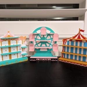 May include: Three carnival-themed wooden display shelves. The left shelf is a carousel with pink and teal accents. The center shelf is a teal and pink Ferris wheel with the text TICKET TO FUN. The right shelf is a red, white, and blue tent.