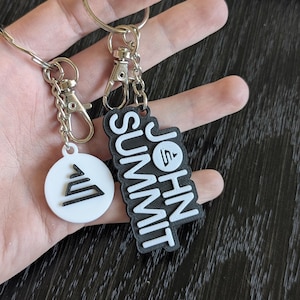 May include: Two keychains with silver-colored hardware. One keychain has a white, circular pendant with a black geometric design. The other keychain has a black, rectangular pendant with the words "JOHN SUMMIT" in white.