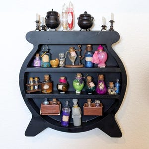 May include: A black cauldron-shaped shelf with three shelves filled with various glass bottles and jars. The top of the shelf has two small black cauldrons and four glass bottles with red and clear liquids.